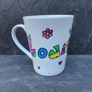 May include: White ceramic mug with a handle, decorated with the name "Lodi" in colourful, playful lettering. The mug features a flower and hearts, adding a whimsical touch. A charming and personalised gift.