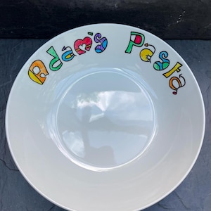 May include: White ceramic pasta bowl with the words "Adam's Pasta" in colourful, hand-painted lettering around the rim. The bowl has a wide, shallow design, perfect for serving pasta dishes. The text includes a heart and other decorative elements.