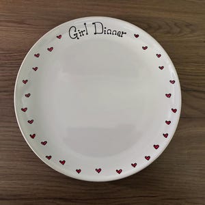 May include: White ceramic plate with the words "Girl Dinner" in black script. The plate is decorated with small red hearts around the rim. The plate is on a wooden surface.