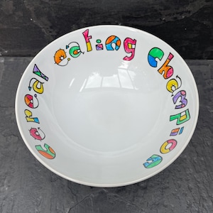 May include: A white ceramic bowl with a colourful hand-painted design that says "Cereal Eating Champion".