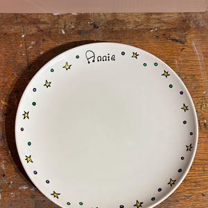 May include: A round white ceramic plate featuring a hand-painted border of small yellow stars and alternating green and purple dots. The name Annie is written in black script at the top centre of the plate, positioned within the decorative border.