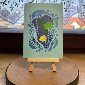 Könnte beinhalten: Eine handgefertigte Grußkarte mit einem geschichteten Papierdesign, das eine Unterwasserszene darstellt. Die Karte zeigt eine grüne Schildkröte, einen gelben Fisch und blaue Wasserpflanzen. Die Karte wird auf einer kleinen Holzstaffelei präsentiert.