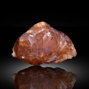 May include: A faceted, amber-colored crystal formation with a reflective surface below. The crystal has sharp edges and a complex, geometric structure. The lighting highlights the crystal's internal structure and color variations.