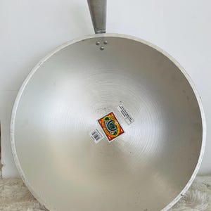 Puede incluir: Un wok grande de aluminio plateado con un mango. El wok tiene una etiqueta en la parte inferior que dice "Hecho en India".