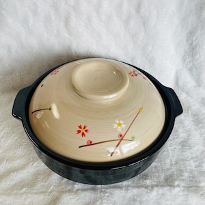May include: A round, black ceramic pot with a matching lid. The lid is beige with a floral design in red, yellow, and white. The pot has two handles and is suitable for cooking.