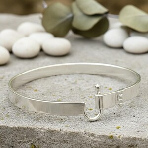 May include: A silver-colored bangle bracelet with a hook and eye closure. The bracelet is made of a flat, polished metal and rests on a gray surface. The clasp features a small hook and a rectangular bar.