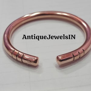 May include: A copper-colored open bangle bracelet. The bracelet has a rounded shape with a gap at the front. The ends of the bracelet are slightly thicker and have etched lines. The text "AntiqueJewelsIN" is printed on the bracelet.