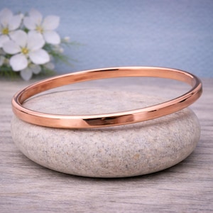 Könnte beinhalten: Ein poliertes roségoldenes Armreif-Armband liegt auf einem glatten, grauen Stein. Das Armband ist ein einfaches, abgerundetes Band. Weiße Blumen und ein hellblauer Hintergrund sind sichtbar.