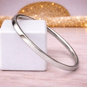 Può includere: Un bracciale rigido d'argento è appoggiato su un cubo bianco contro uno sfondo rosa e oro. Il bracciale è una fascia semplice e lucida, che riflette la luce. Questo gioiello è un accessorio classico.