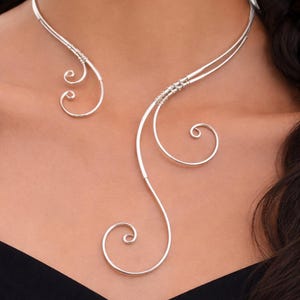 May include: A silver-toned necklace with a unique, flowing design. The necklace features two curved lines that meet at the back, with delicate spiral accents. The jewelry is worn against a dark background.
