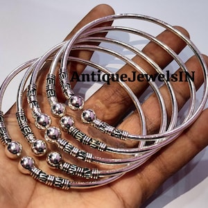 Op de afbeelding: Een verzameling zilverkleurige armbanden. Elke armband heeft een gladde, afgeronde band met decoratieve details aan de uiteinden, waaronder een kleine zilveren bal. De armbanden worden op een hand getoond. De tekst "AntiqueJewelsIN" is zichtbaar.