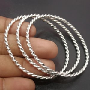 Könnte beinhalten: Vier silberfarbene Armreifen mit einem gedrehten Seildesign. Die runden Armreifen scheinen aus Metall zu sein. Der Schmuck wird vor einem dunklen Hintergrund präsentiert.