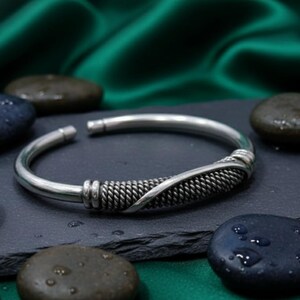 Könnte beinhalten: Ein silberfarbenes Armreif-Armband mit einem gewebten Design in der Mitte. Das offene Armband liegt auf einer dunkelgrauen Oberfläche, umgeben von glatten, dunklen Steinen. Der Hintergrund zeigt einen grünen, seidigen Stoff.