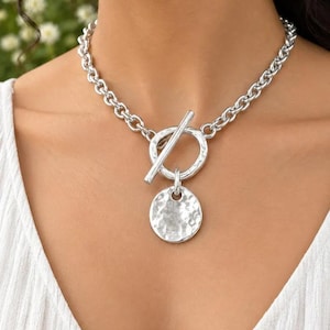 Puede incluir: Un collar de cadena en tono plateado con un cierre de barra en la parte delantera. El collar incluye un componente de anillo circular y un colgante redondo con textura martillada que cuelga del mecanismo de cierre, con acabado metálico.