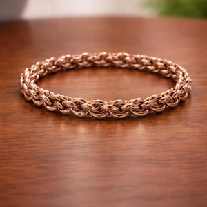 Peut inclure: Un bracelet en or rose avec un motif de chaîne tissée. Le bracelet est circulaire et repose sur une surface en bois. Les tons chauds du métal contrastent avec le grain du bois.