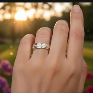 May include: A silver ring with a wave-like design is worn on a finger. The ring has a polished finish and a detailed pattern. The background is blurred, with sunlight and greenery visible.