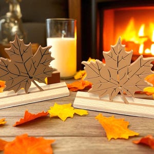 Könnte beinhalten: Zwei Ahornblatt-Dekorationen aus Holz mit braunen Blattdetails und hellen Holzsockeln. Die Blätter liegen auf einer Holzoberfläche, die mit bunten Herbstblättern bedeckt ist, mit einem Kamin und einer Kerze im Hintergrund.