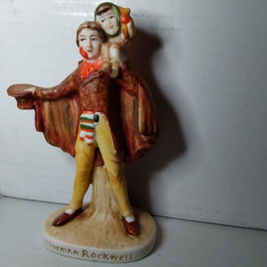 Puede incluir: Una figura de cerámica que representa a un hombre con capa marrón y pantalones amarillos, llevando a un niño sobre sus hombros. La figura tiene una bufanda roja y un sombrero. La base es blanca con el texto "Norman Rockwell".