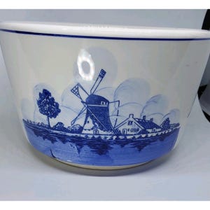 May include: A white ceramic mixing bowl with a blue Dutch windmill and landscape design. The bowl has a wide rim and a curved shape, with the blue design wrapping around the lower portion of the bowl.