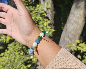 May include: A beaded bracelet featuring a mix of blue, green, white, and yellow beads. The bracelet includes a gold-colored charm and is worn on a wrist with a beige sweater sleeve. The background is a blurred view of a tree.