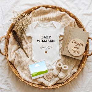 May include: A white ribbed baby bodysuit with the text BABY WILLIAMS and April 2027 printed in black, featuring a small brown heart. Includes a pair of cream knit booties, two books, a wooden disc reading Chapter Two Begins, and a feather.