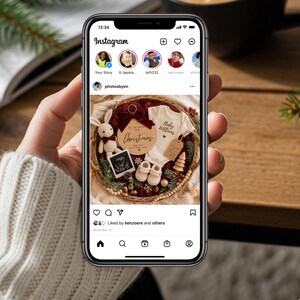 May include: A smartphone screen displays a festive baby announcement. The arrangement includes a white onesie, baby shoes, a wooden Christmas sign, a stuffed bunny, and a sonogram image, all within a woven basket. The text on the sign reads "Christmas" and "Baby Williams".