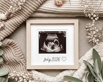 May include: A framed ultrasound image with a baby's profile is displayed. The frame is light wood, and the image is surrounded by a white mat. The text "July 2026" is written below the image, with a heart symbol. The background includes a beige knitted blanket and decorative flowers.