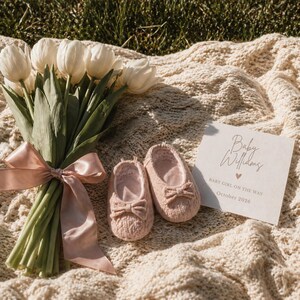 May include: A bunch of white tulips with a pink ribbon, beside pink baby shoes with bows. A card reads "Baby Williams, Baby Girl on the Way, October 2026". The items are arranged on a cream knitted blanket. A baby announcement.