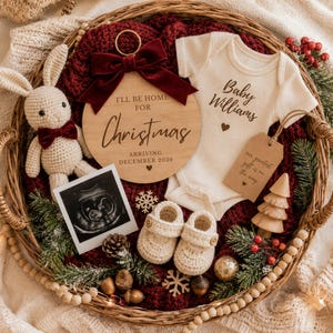 May include: A festive baby announcement in a woven basket. Includes a wooden ornament with "I'll be home for Christmas" text, a baby onesie, ultrasound photo, crocheted booties, and a plush bunny. The theme is a Christmas baby arrival.