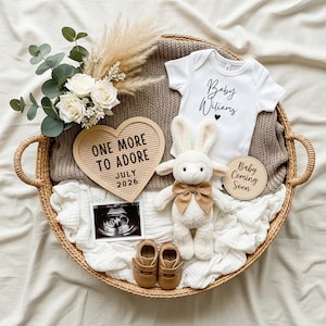 Może przedstawiać: Wiklinowy kosz zawiera elementy ogłoszenia o dziecku. Białe body z napisem "Baby Williams", pluszowa owieczka, tabliczka w kształcie serca z napisem "ONE MORE TO ADORE JULY 2026", buciki dla dziecka i USG ułożone z kwiatami.