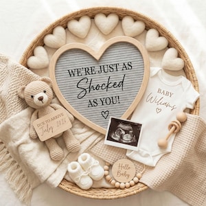 Può includere: Un cesto di vimini contiene articoli per l'annuncio del bambino. Una lavagna a forma di cuore grigio recita "WE'RE JUST AS SHOCKED AS YOU!". Un body bianco dice "BABY Wiliams". Un orsacchiotto all'uncinetto tiene un cartello: "DUE TO ARRIVE July 2026".