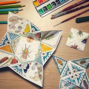 May include: Two origami paper stars with a variety of colorful insects and designs. The stars are folded from paper with blue, green, yellow, and red accents. The stars are on a wooden surface with a watercolor paint set and paintbrushes.