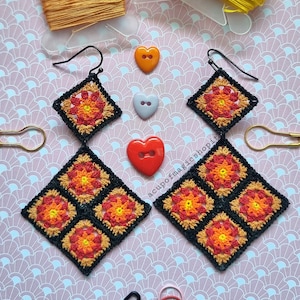 Può includere: Orecchini all'uncinetto con bordo nero e design floreale arancione, giallo e rosso. Gli orecchini quadrati hanno una chiusura a gancio. L'immagine include anche bottoni a forma di cuore, filo e altri materiali per l'artigianato.