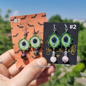 Könnte beinhalten: Zwei Paar handgefertigte Ohrringe. Ein Paar hat ein grünes, gelbes und schwarzes Häkeldesign mit einem dunkelvioletten und grünen Stein. Das andere Paar hat ein ähnliches Häkeldesign mit rosa Perlen. Der Text "acupofmagicshopit" ist sichtbar.