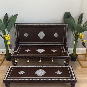 May include: Dark brown wooden bench and footstool set with intricate silver inlay designs. The bench has a backrest and is flanked by potted plants with large green leaves and yellow fruit. The set is placed in front of a gold arch.