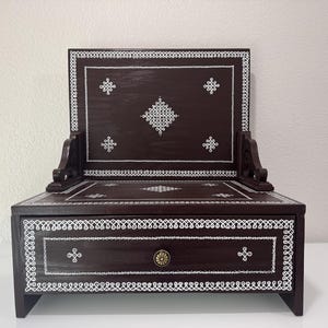 May include: A dark brown wooden decorative box with a hinged back and a drawer. The box is adorned with white, intricate, geometric patterns and a brass knob. The design is symmetrical and the box is set on four short legs.