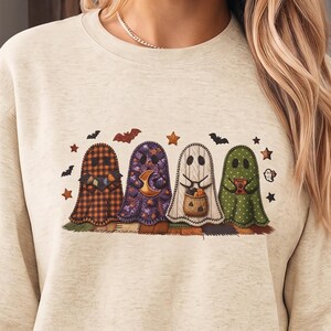 Peut inclure: Un sweat-shirt à col rond beige avec une impression graphique représentant quatre fantômes alignés. Les fantômes présentent différents motifs, notamment un tartan orange, des fleurs violettes, des rayures blanches et des pois verts, avec des chauves-souris et des étoiles.