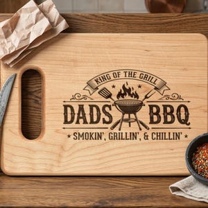 May include: A wooden chopping board with a handle, engraved with "KING OF THE GRILL" and "DADS BBQ". Features a barbecue graphic and the text "SMOKIN', GRILLIN', & CHILLIN'". A fork, knife, and a bowl of spices are on the wooden surface.