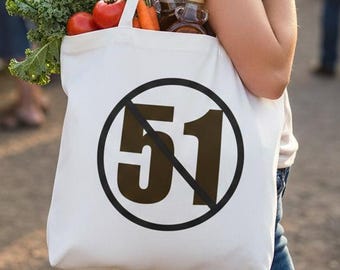 May include: A white canvas tote bag featuring a black circular emblem with a diagonal line through the number 51. The bag is filled with fresh produce including a loaf of bread, green kale, red tomatoes, orange carrots, and two small glass bottles.