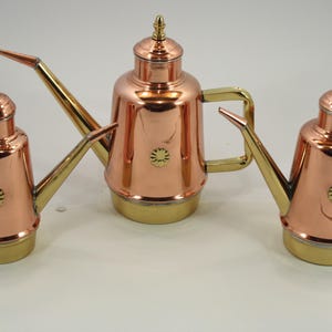 Può includere: Tre versatori d'olio in rame e ottone. Ognuno ha un corpo in rame, un beccuccio, un manico e una base in ottone. I versatori hanno un emblema floreale decorativo sul davanti e un piccolo pinnacolo sul coperchio.
