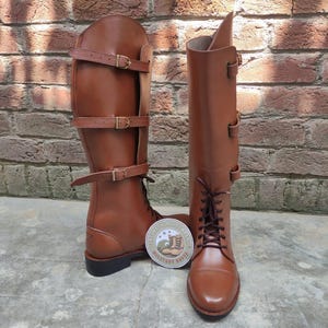 May include: A pair of brown leather riding boots with multiple buckle straps. The boots have a tall shaft and a lace-up design at the ankle. A round sticker with text and a crest is placed between the boots.