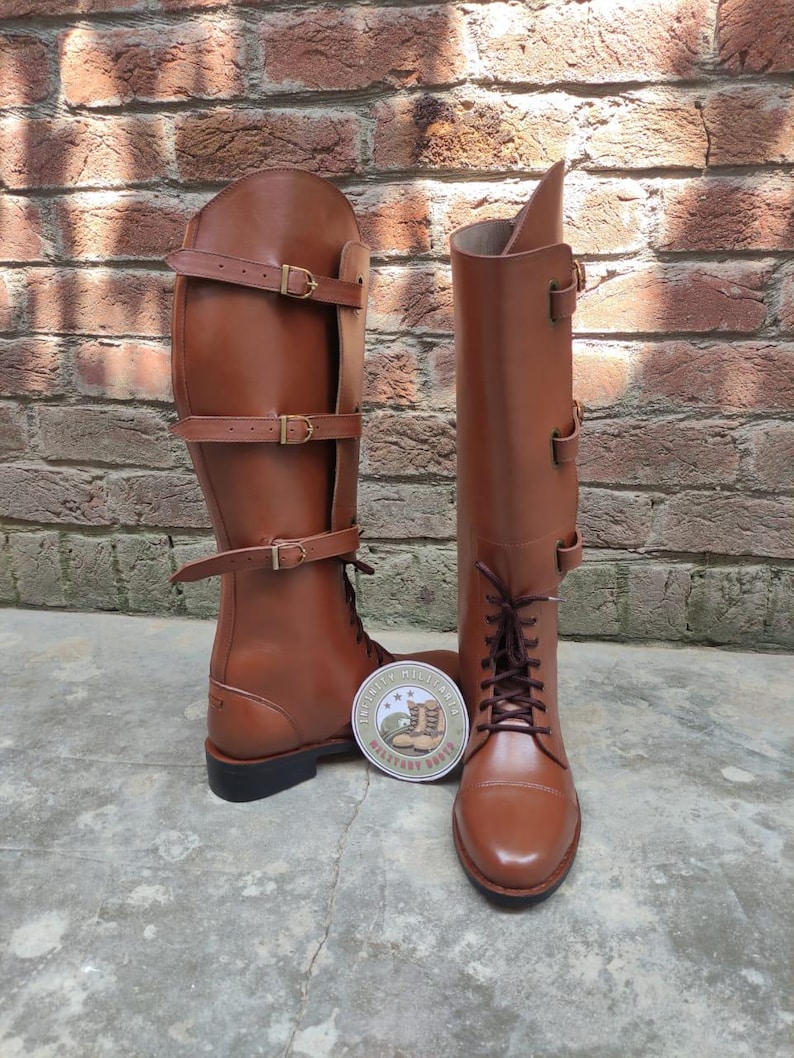 May include: A pair of brown leather riding boots with multiple buckle straps. The boots have a tall shaft and a lace-up design at the ankle. A round sticker with text and a crest is placed between the boots.