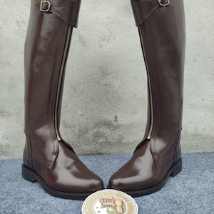 May include: A pair of tall, brown leather riding boots with gold zippers and buckles. The boots have a classic design, with a smooth, polished finish. The boots are displayed on a blue surface, with a circular logo in the foreground.