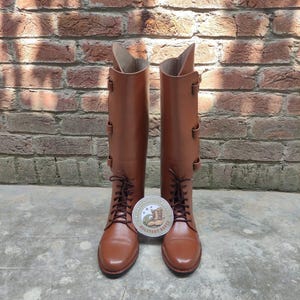 May include: A pair of brown leather riding boots with laces and buckles. The boots are tall, reaching the knee, and are set against a brick wall background. A round sticker with text and a boot illustration is between the boots.