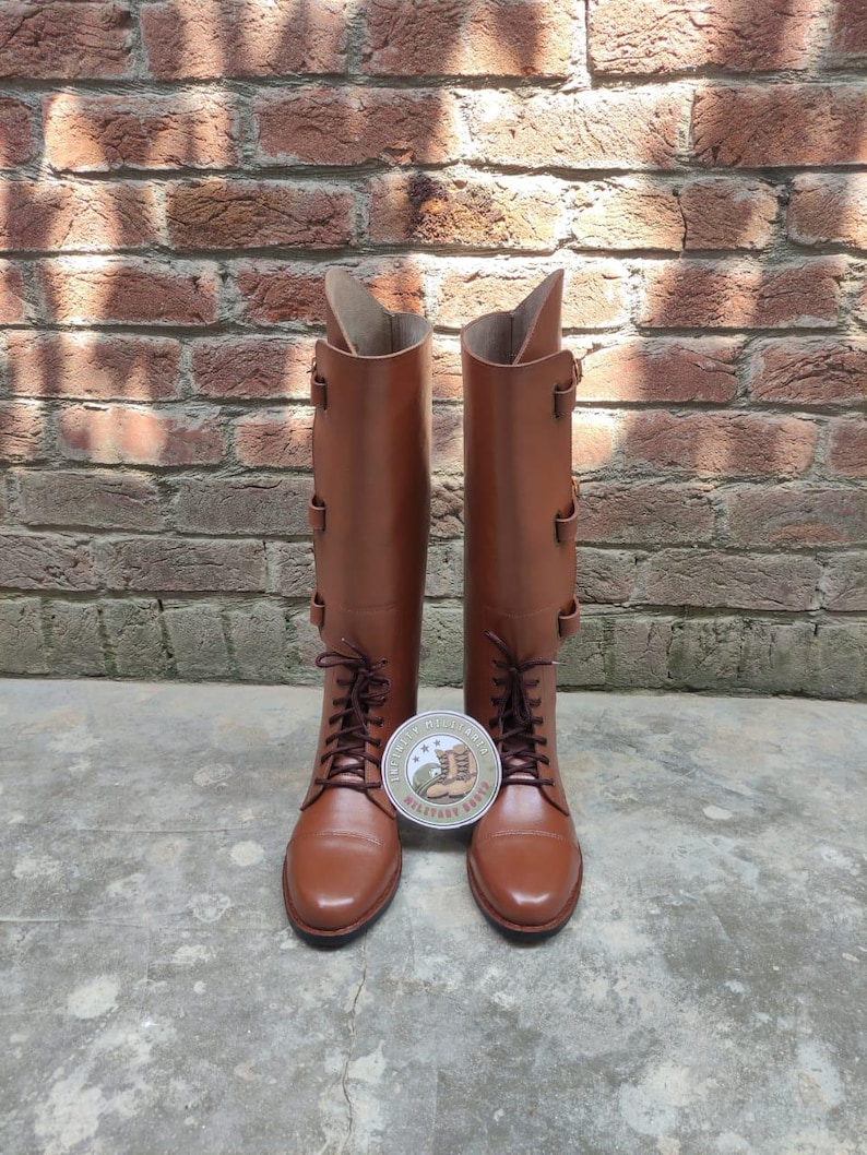 May include: A pair of brown leather riding boots with laces and buckles. The boots are tall, reaching the knee, and are set against a brick wall background. A round sticker with text and a boot illustration is between the boots.