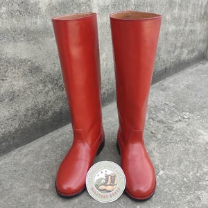 Puede incluir: Un par de botas altas de cuero rojo. Las botas son hasta la rodilla con un acabado liso y brillante. Una pegatina circular con el texto "INFINITY MILITARIA MILITARY BOOTS" se coloca entre las botas. Estilo ecuestre clásico.