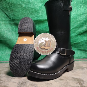 Puede incluir: Botas de cuero negro con una correa de hebilla y punta redonda. Las botas tienen una suela gruesa y un tacón apilado. Se ve la suela de una bota, que muestra la talla 6. Una etiqueta circular dice "INFINITY MILITARIA BOOTS".