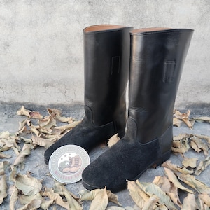 Puede incluir: Un par de botas de cuero negro con secciones inferiores de gamuza. Las botas tienen una caña alta y una puntera redonda. Una pegatina circular con texto y un gráfico de bota es visible entre las botas. Hojas secas rodean las botas.
