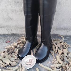 May include: A pair of tall, black leather boots with a smooth, glossy finish. The boots have a classic design, with a high shaft and a rounded toe. A circular emblem with text and a boot illustration is visible.