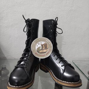 Puede incluir: Par de botas militares de cuero negro con cordones. Las botas tienen un parche redondo con el texto "INFINITY MILITARIA BOOTS". Las suelas son marrones con un detalle de costura blanca. Las botas se exhiben sobre una superficie de vidrio.